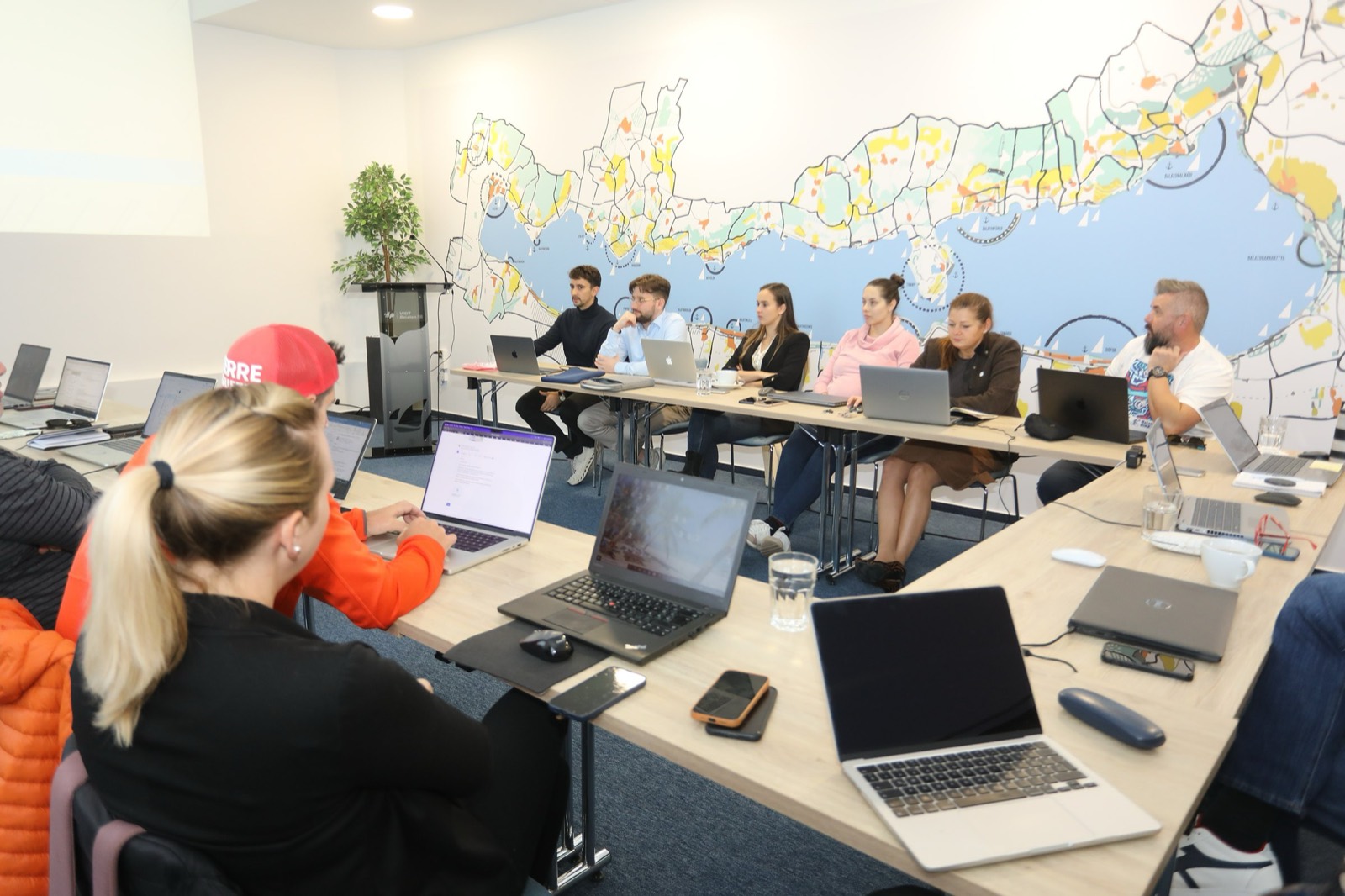 01: AI training in the VisitBalaton365 training room in Keszthely; participants working on laptops, the AI slide on the screen, the map of Lake Balaton on the wall. 02: VisitBalaton365 DMO staff at the U-shaped tables during the AI training, in front of the Lake Balaton map wall. 03: Participants practicing AI tools on laptops during the training. 04: The full group of the training room at work. 05: DMO staff attentively following the AI training demo.