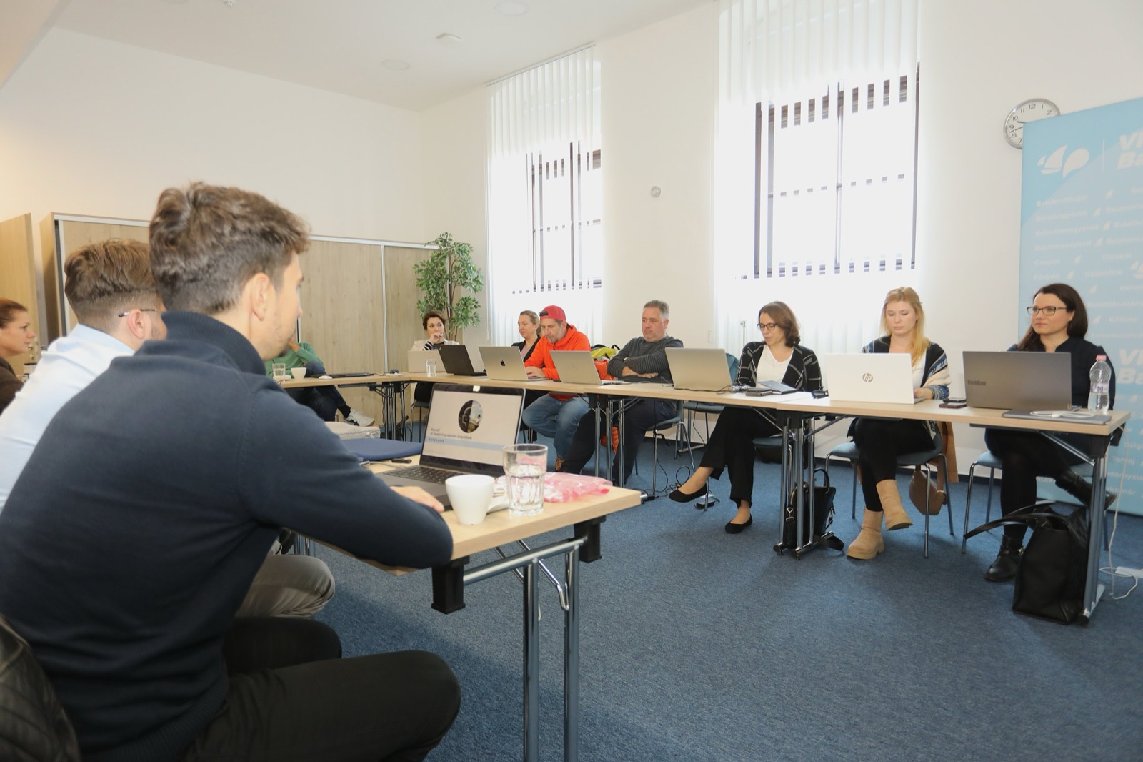 01: AI training in the VisitBalaton365 training room in Keszthely; participants working on laptops, the AI slide on the screen, the map of Lake Balaton on the wall. 02: VisitBalaton365 DMO staff at the U-shaped tables during the AI training, in front of the Lake Balaton map wall. 03: Participants practicing AI tools on laptops during the training. 04: The full group of the training room at work. 05: DMO staff attentively following the AI training demo.