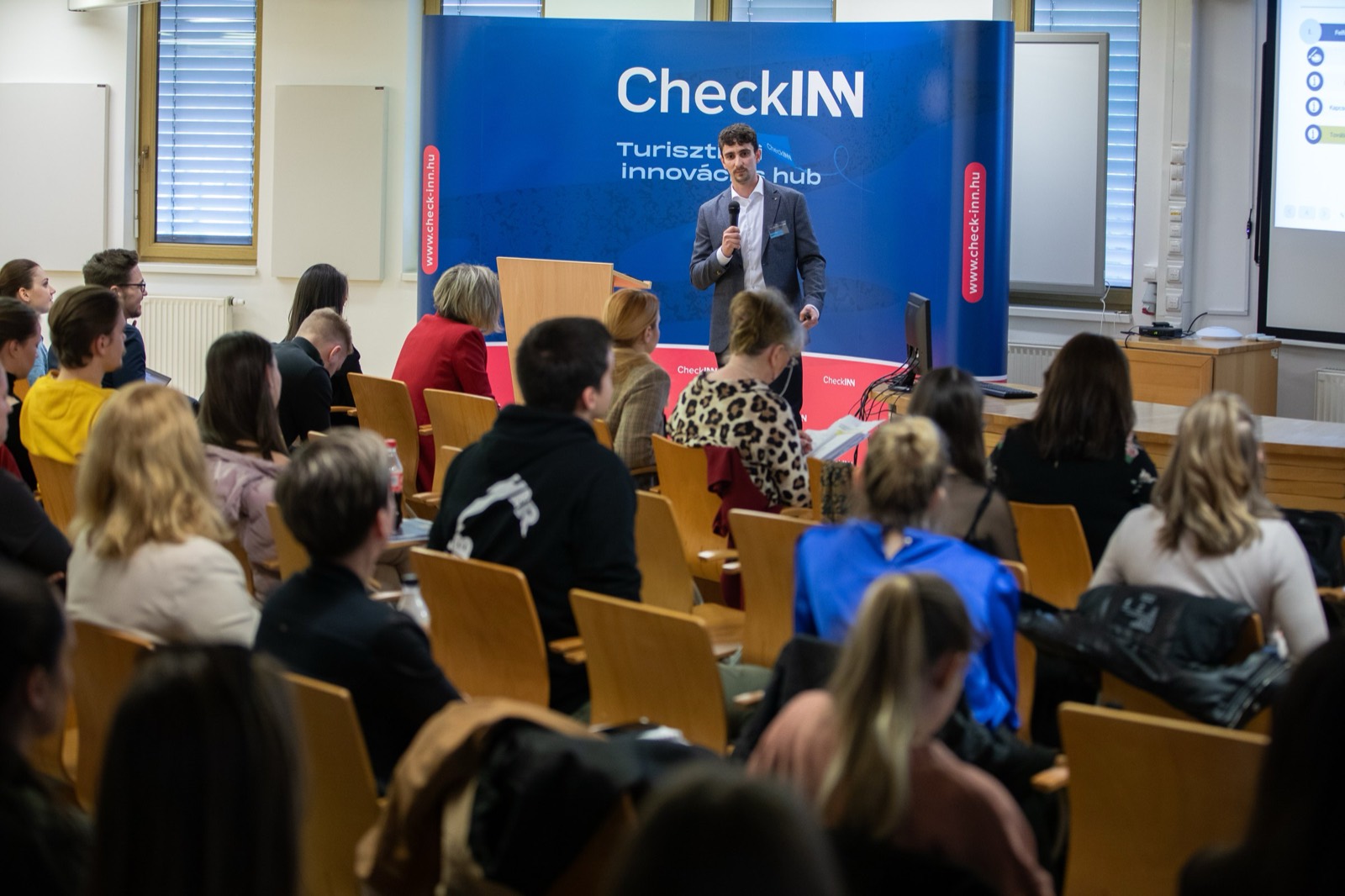 01: Adam Schmutz speaking with a microphone in front of the CheckINN banner, with the audience in the foreground. 02: Adam Schmutz mid-talk, the CheckINN Tourism Innovation Hub banner in the background. 03: Adam Schmutz presenting to the workshop audience in front of the CheckINN backdrop. 04: The full lecture hall with the audience; Adam Schmutz on stage by the CheckINN banner. 05: Round-table discussion: Adam Schmutz and the moderator in front of the METU backdrop. 06: The participants of the workshop's round-table discussion in front of the Budapest Metropolitan University banner. 07: Adam Schmutz during a professional conversation in a break of the event.